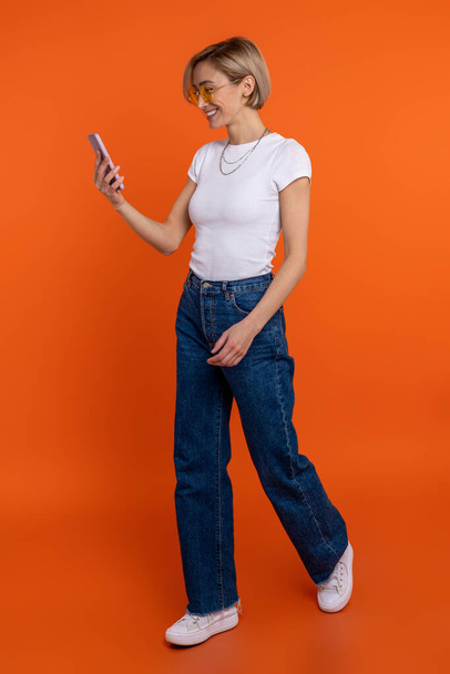Woman blogger in white t shirt and jeans using smart phone browsing internet isolated over orange background. - Foto, immagini
