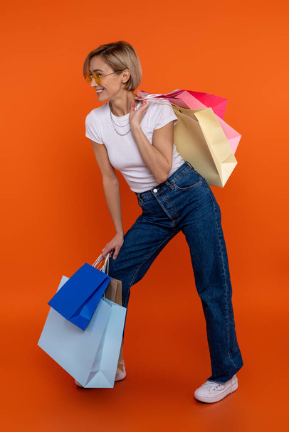 Delighted woman in white t shirt and jeans enjoying shopping holding bags isolated over orange background. - Foto, Imagem