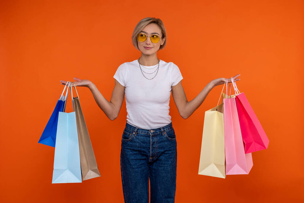 Pensive woman in white t shirt and jeans holding shopping bags in both hands isolated over orange background. - Фото, изображение