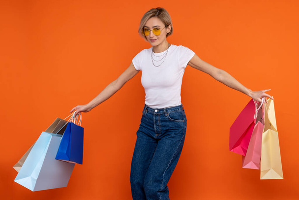 Festive woman in white t shirt and jeans holding bags enjoying shopping isolated over orange background. - Foto, Imagem