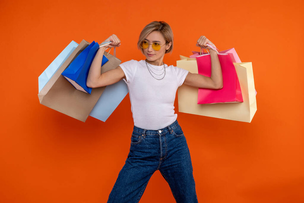 Funny strong woman in white t shirt and jeans raised arms with shopping bags isolated over orange background. - Valokuva, kuva