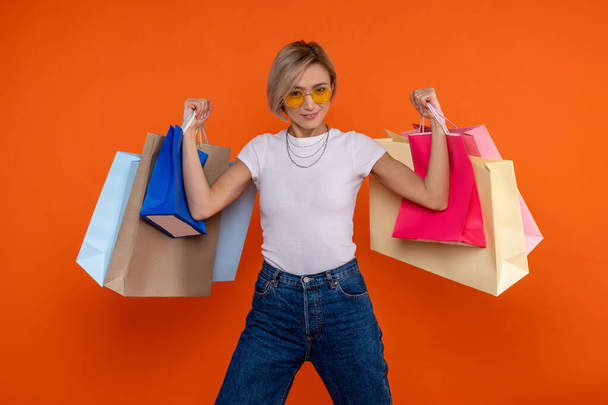 Funny strong woman in white t shirt and jeans raised arms with shopping bags isolated over orange background. - Photo, Image