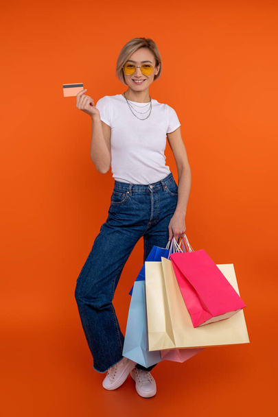 Satisfied woman in white t shirt and jeans holding shopping bags and credit card isolated over orange background. - Fotografie, Obrázek