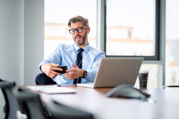 Portrait of businessman using smartphone and earphones while sitting at the office and having a video call. Professional man wearing shirt and tie.  - Foto, Imagem