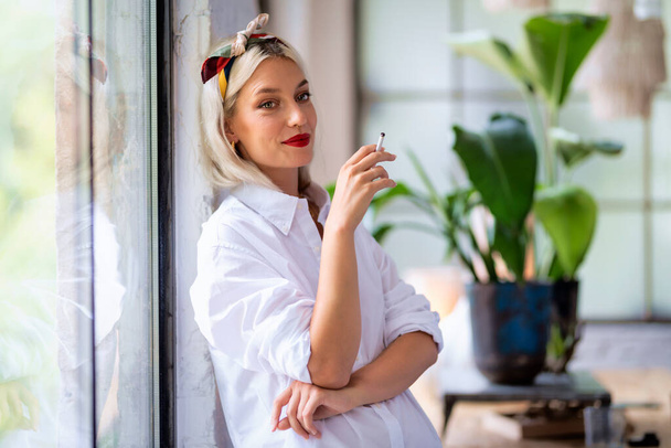 Portrait shot of beautiful young woman wearing hair scarf and white shirt while smoking cigarette by the window at home. - Фото, изображение
