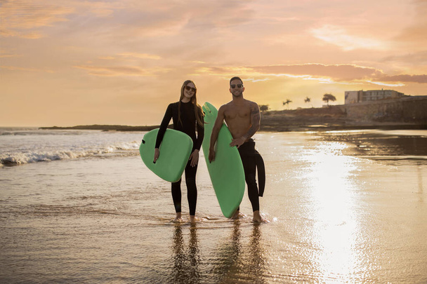 Молодые серферы в паре с досками для сёрфинга в руках, гуляющие по пляжу на закате, счастливые улыбающиеся друзья Мужчина и женщина веселятся вместе, наслаждаясь серфингом и активным образом жизни, копировать пространство - Фото, изображение