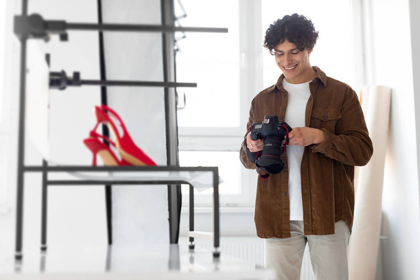 Mutlu profesyonel fotoğrafçı modern fotoğraf makinesinde fotoğrafları kontrol ederken içerik fotoshoot yapıyor, kırmızı şık ayakkabıların yanında duruyor. - Fotoğraf, Görsel
