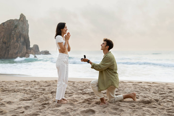 Romanttinen ehdotus meren rannalla. Rakastava nuori mies kihlasormus tekee ehdotuksen onnellinen nainen rannalla rantaviivalla, sivunäkymä - Valokuva, kuva