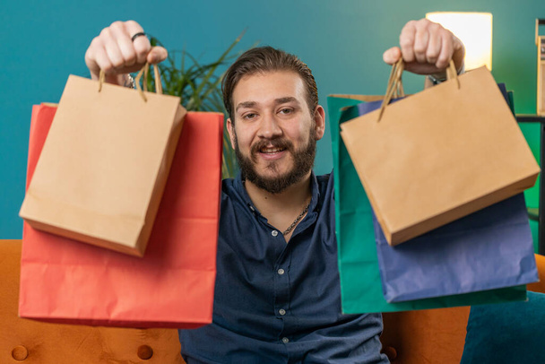 Young lebanese man happy shopaholic consumer came back home after shopping sale with bags. Portrait of arabian guy satisfied received parcels purchase from online order at home apartment room on sofa - Photo, Image