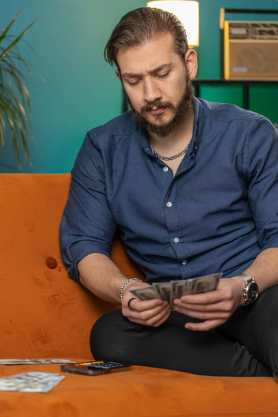 Planungsbudget. Reicher libanesischer Mann, der Geld in bar zählt, nutzt Smartphone-Taschenrechner-App, um Rechnungen zu Hause zu berechnen. Araber zufrieden mit Einkommen, Gewinn, spart Geld für geplanten Urlaub - Foto, Bild
