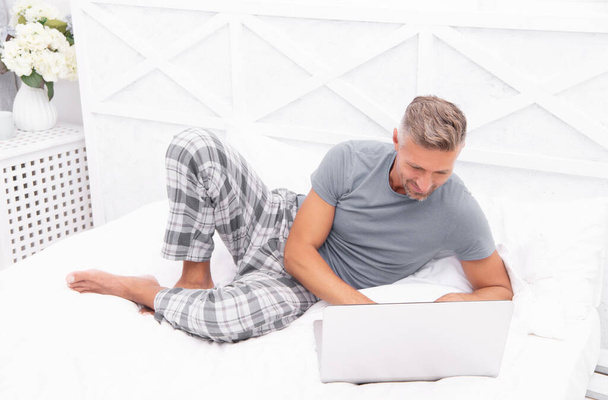 positive man freelancer at home in bed. man freelancer at home in bedroom. photo of man freelancer at home with laptop. man freelancer at home. - Fotó, kép