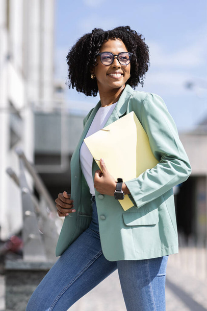 Vertical outdoor shot of beautiful millennial african american business woman going to office, business meeting, looking aside and smiling. Happy pretty black lady in smart casual outfit, eyeglasses - Fotoğraf, Görsel