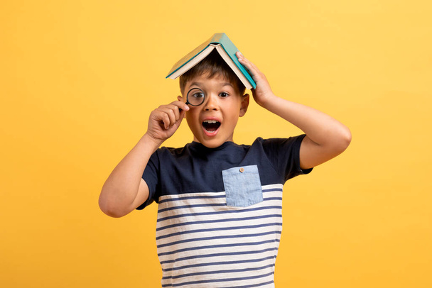 Excited curious kid cute school aged boy with book on his head holding magnifier, child bookworm getting interesting knowledge while reading, isolated on yellow studio background - Фото, изображение