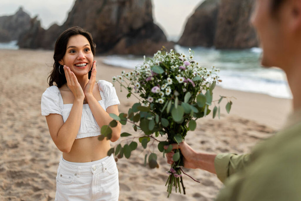 Glückliches erstaunliches Paar mit Date oder Heiratsantrag am Strand, liebevoller Freund gibt Blumenstrauß an überraschte Freundin, feiert Jubiläum - Foto, Bild