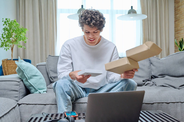 Joven hombre desempacar una compra en línea, sentado en el sofá en casa con caja con nuevo teléfono inteligente. Internet compras en línea, entrega, concepto de juventud - Foto, imagen