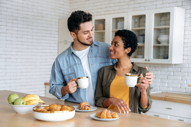 Koffie tijd in de ochtend. Gelukkige echtgenoten van verschillende nationaliteiten hebben lekker weekend ontbijt in de keuken thuis met croissants en koffie, samen tijd doorbrengen, naar elkaar kijken, glimlachen - Foto, afbeelding