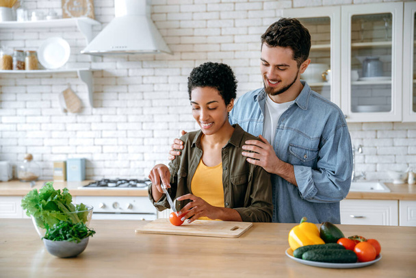 Glückliches multiethnisches Ehepaar, in Freizeitkleidung, junge Ehepartner, stehen zu Hause in der Küche, bereiten Salat zu, Frau schneidet Gemüse, Mann umarmt sie, sie freuen sich, Zeit miteinander zu verbringen, lächeln - Foto, Bild