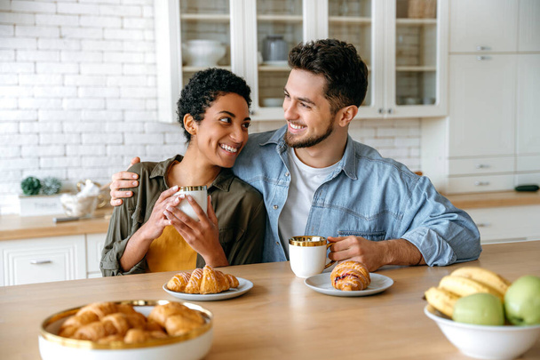 Heureux jeune couple familial, homme caucasien et femme afro-américaine, prenant le petit déjeuner à la maison confortable cuisine avec croissants et café, regardez-vous, sourire. Dimanche matin ensemble - Photo, image