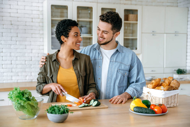 Happy married couple in casual wear, young spouses, stand at home in the kitchen, prepare fresh salad together, woman cuts vegetables, man hugs her, they look at each other with love, smile - Photo, Image