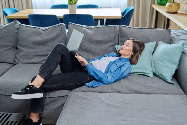 Young woman lying on sofa using laptop. Internet technologies for study leisure, female freelancer working remotely, university student watching online course, rest communication in social networks - Foto, Imagem