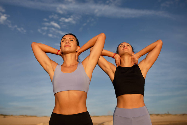 Cheerful calm young european women twins in sportswear doing hand stretching, enjoy morning yoga at sea beach, outdoor. Summer workout together, active lifestyle, body care and weight loss - Foto, Imagen