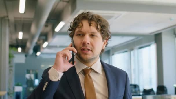 Borstfoto van een blanke zakenman in een formeel pak die door kantoor loopt, mobiele telefoon praat en knikt met zijn hoofd om collega te begroeten - Video