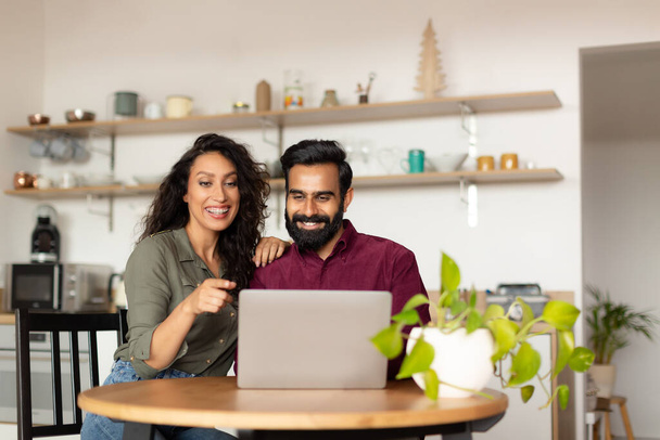 Happy arab spouses paying taxes online with laptop in kitchen, couple using computer, planning family budget or shopping online together, free space - Fotó, kép
