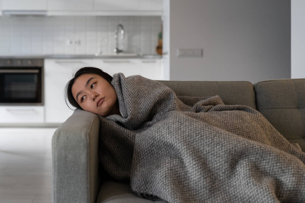 Inintéressé fatigué asiatique coréen jeune femme regarde tristement loin de mauvaise humeur se trouve sur le canapé sous couverture de laine à la maison pense à des problèmes de dépression dans la santé mentale, burnout ou le cœur brisé - Photo, image