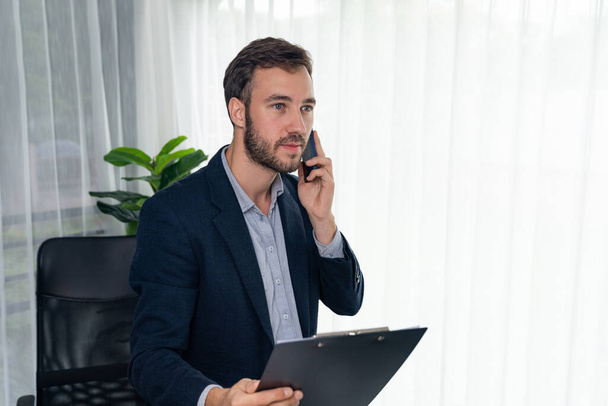 Hardwerkende en ijverige zakenman werkt aan zijn laptop terwijl hij praat met klanten op zijn mobiele telefoon, presentatie van moderne multitasking kantoormedewerker levensstijl. Entiteit - Foto, afbeelding