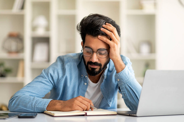 Young Indian Freelancer Man Checking Working Schedule In Notepad At Home, Handsome Eastern Male Sitting At Desk Indoors, Looking At Planner Notebook, Managing His Timeline, Free Space - Foto, afbeelding