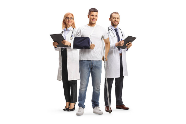 Young man with crutches and arm sling standing in front of a team of doctors isolated on white background - Foto, imagen