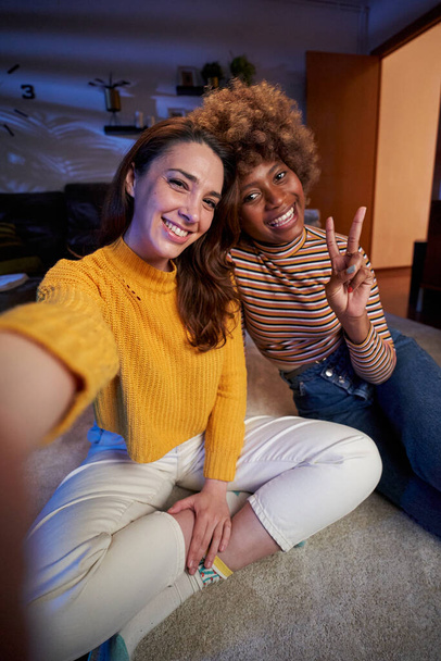 Vertical in love LGBT multiracial young lesbian couple phone selfie posing smiling to camera. Portrait two cheerful women together sitting on carpet living room at night. People relationship at home. - Foto, afbeelding