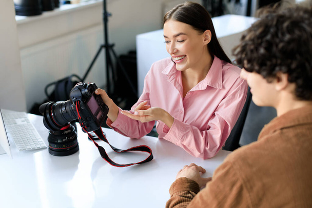 Happy woman photographer showing photos after shooting, discussing work with man in studio, talking after photoshoot and choosing the best pictures, sitting at workplace - Foto, Imagem