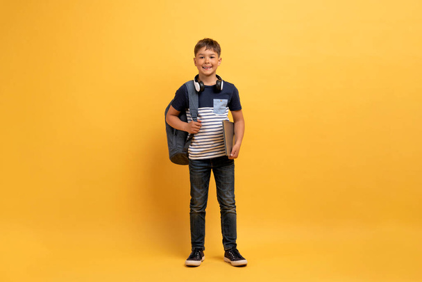Studio shot of cheerful cute handsome caucasian boy wearing casual outfit, carrying backpack and books smiling at camera, schooler posing on yellow background, full length, copy space - Foto, Imagen