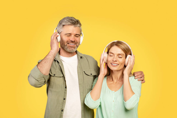 Gelukkig Europees echtpaar van middelbare leeftijd met een koptelefoon en luisterend naar muziek met gesloten ogen, man knuffelende vrouw, poserend over gele achtergrond. Echtgenoten genieten van favoriete sountracks - Foto, afbeelding