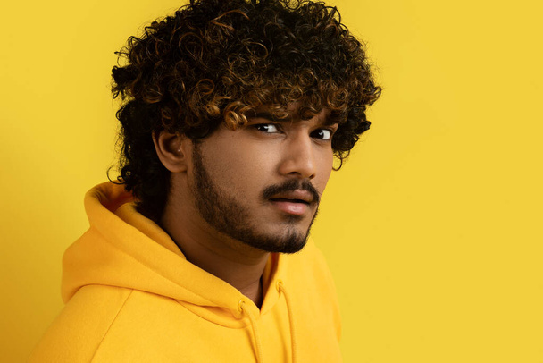 Portrait of unsure young indian bearded curly man 30s with suspicious look wearing yellow hoodie posing on camera with raising eyebrow isolated over yellow background, closeup, copy space - 写真・画像