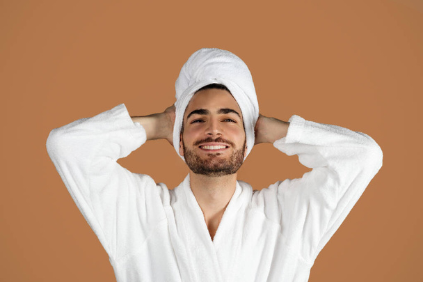 Beauty Spa Day. Happy Bearded Arabic Young Man Posing With White Bathrobe And Towel, Holding Hands Behind Head, Relaxing And Enjoying Self Care Routine On Brown Background. Studio Portrait - Fotografie, Obrázek