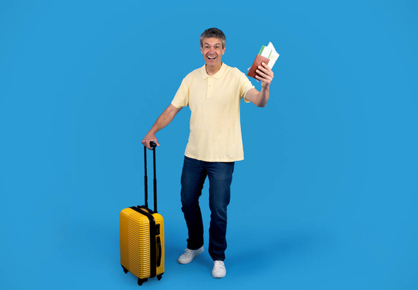 Reiseangebot. Grauer Touristenmann mit Koffer posiert und zeigt vor Freude über den blauen Studiohintergrund seine Bordkarte. Schuss glücklicher Weltenbummler mit Gepäck und Tickets bereit für die Urlaubsreise - Foto, Bild