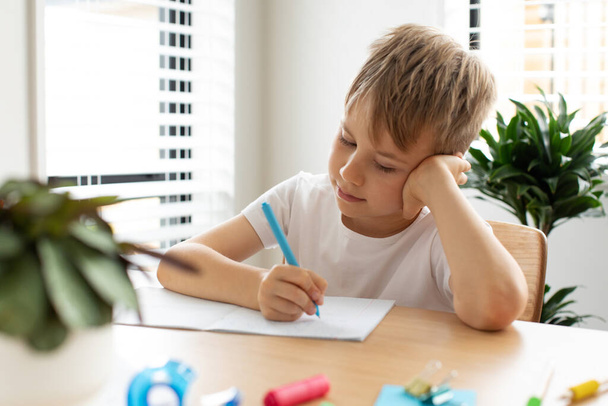 Cute boy doing school homework at home. Distance education. Online education. The boy is writing intently in a notebook. - Fotografie, Obrázek