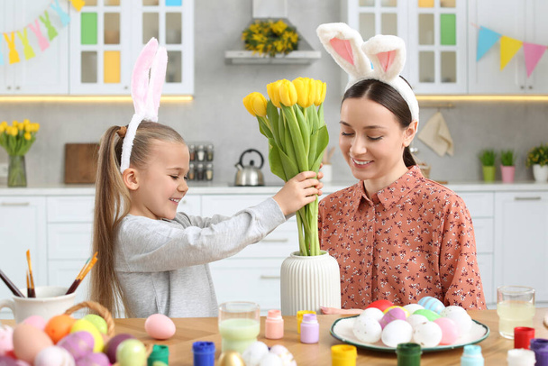 Mother with her cute daughter putting yellow tulips into vase near beautifully painted Easter eggs at table in kitchen - Valokuva, kuva