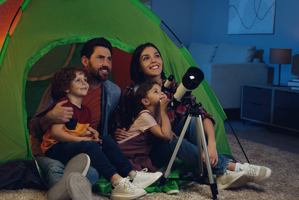 Happy family using telescope to look at stars while sitting in camping tent indoors - Фото, зображення