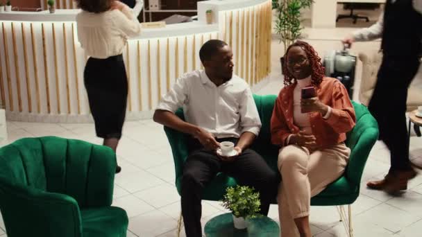 Happy husband and wife in video conference with mates during hotel stay. Relaxed couple on their honeymoon trip in video call with friends while enjoying cup of coffee in resort lobby - Footage, Video