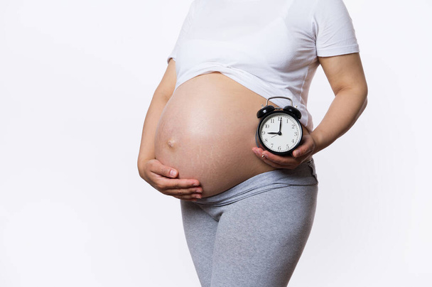Koncept termínu pro miminko. Blízko břicha těhotné ženy, vyčkávající matka držící budík poblíž nahého břicha, izolované na bílém pozadí. Porod. Těhotenství. Čekání na dítě - Fotografie, Obrázek