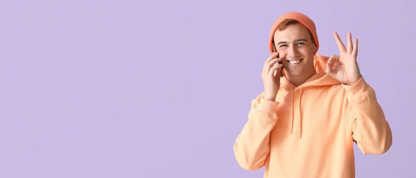 Młody człowiek z telefonem komórkowym pokazuje OK gest na liliowym tle z miejscem na tekst - Zdjęcie, obraz