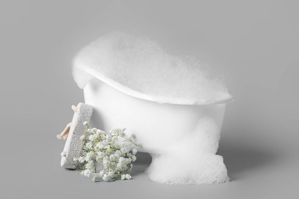 Piccola vasca da bagno con schiuma, fiori di gypsophila e pietra pomice su sfondo grigio - Foto, immagini