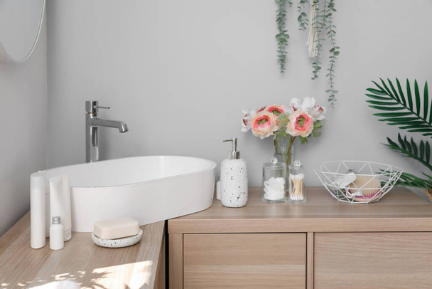 Sink bowl, bath accessories and ranunculus flowers on wooden cabinet in light bathroom - Valokuva, kuva