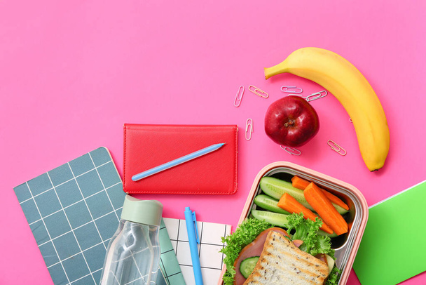 Papeterie différente, boisson et boîte à lunch avec des aliments savoureux sur fond rose - Photo, image