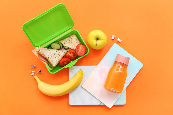 Stationery, drink and lunch box with tasty food on orange background - Photo, Image