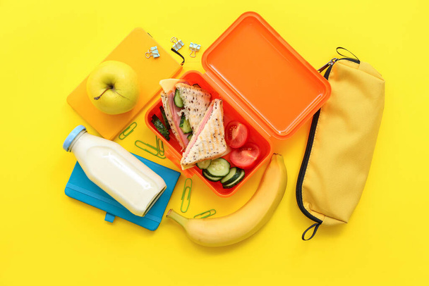 Διάφορα γραφική ύλη, ποτό και μεσημεριανό κουτί με νόστιμο φαγητό σε κίτρινο φόντο - Φωτογραφία, εικόνα