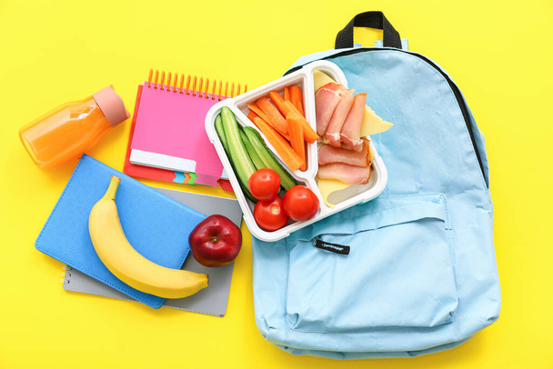 Рюкзак, канцелярские принадлежности, напитки и обеденная коробка с вкусной едой на желтом фоне - Фото, изображение